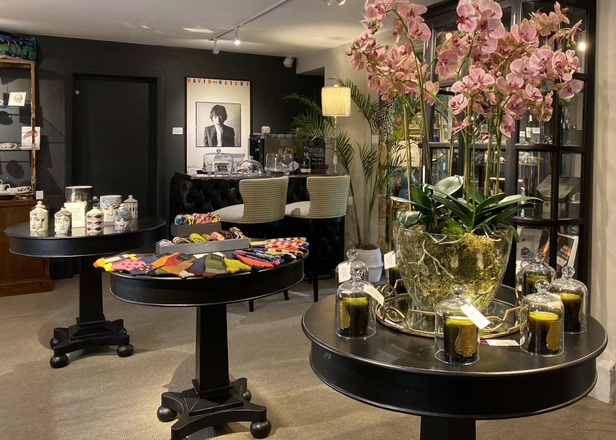 Interior of Barnbury, a stylish independent shop in Winchcombe. In the foreground, a large glass planter filled with pink orchids sits on a round black display table surrounded by decorative candles. Nearby tables display colorful patterned socks and home interior items. In the back, two upholstered chairs sit at a small counter beneath a black-and-white portrait poster, with shelves of home décor and plants throughout the space.
