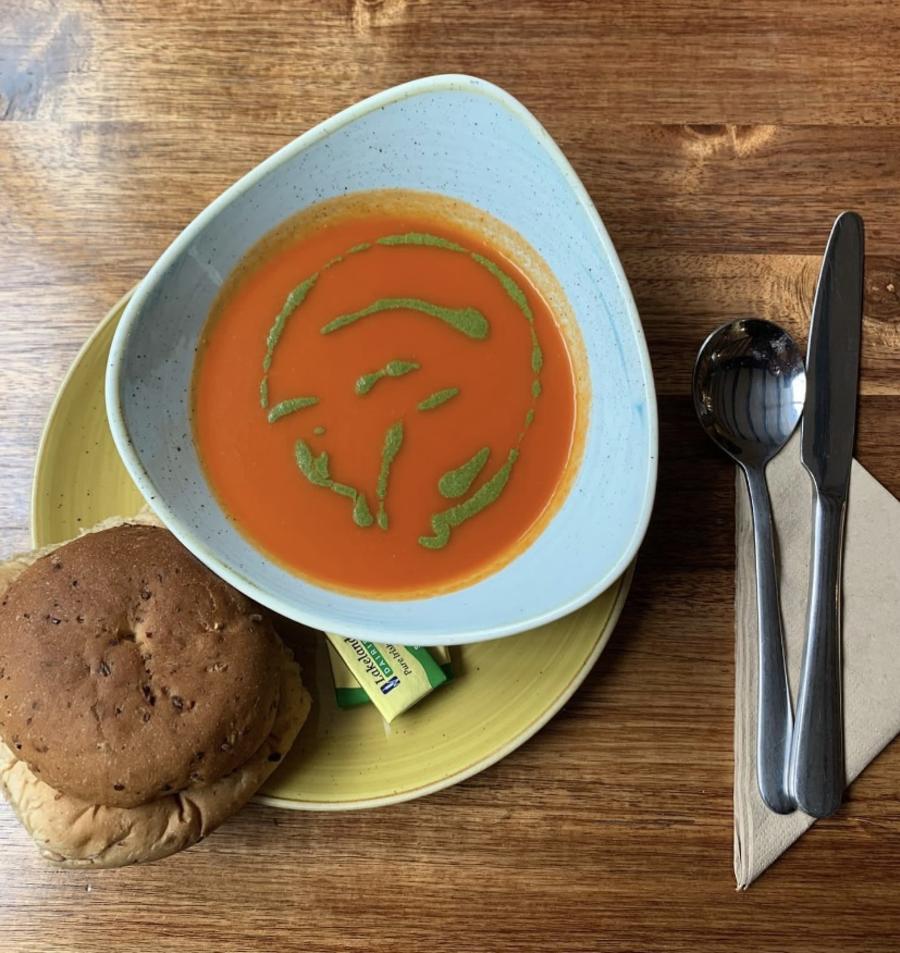 Overhead view of a place setting on a wooden table featuring a bowl of smooth tomato soup drizzled with green herb oil. The bowl sits on a light green plate, accompanied by a bread roll. To the right are a neatly placed knife and fork on a folded napkin, along with a small condiment container. The scene is warmly lit, highlighting the textures of the wood and the vibrant color of the soup.
