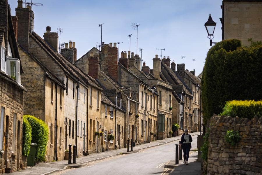A narrow, gently curving street lined with closely packed stone houses featuring sloped roofs, chimney stacks, and small windows. The buildings vary in warm neutral tones like beige, cream, and light brown, with some greenery climbing walls and hedges along the edges. A single pedestrian walks along the pavement on the right side of the road. A low stone wall and neatly trimmed bush appear in the foreground, while traditional street lamps rise above the homes. The sky overhead is pale and clear, casting soft daylight across the quiet scene in Winchcombe.
