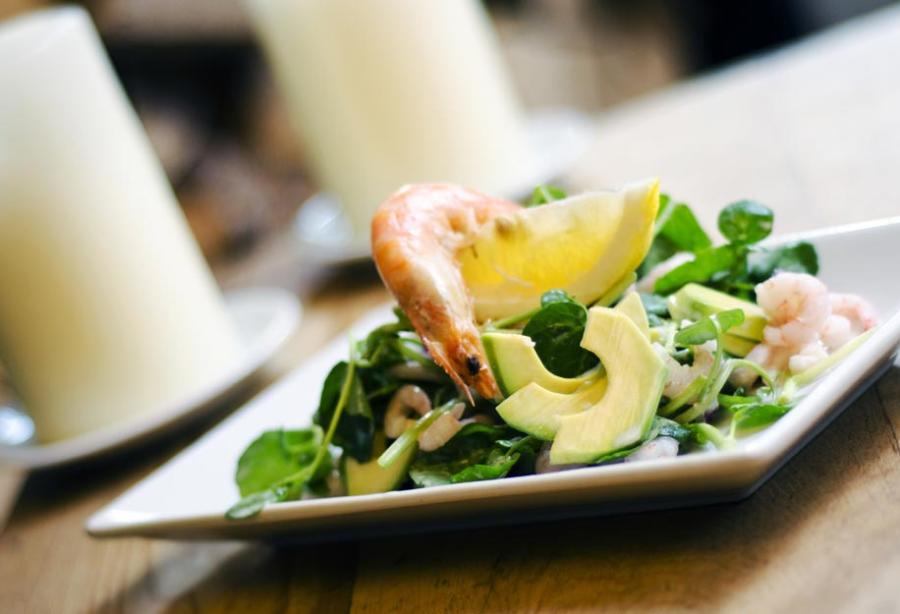White rectangular plate holding a composed salad with sliced avocado, leafy greens, and pieces of seafood, accompanied by a lemon wedge. The dish is placed on a wooden table, with a softly blurred background featuring a white candle and dining setting, creating a warm restaurant atmosphere.