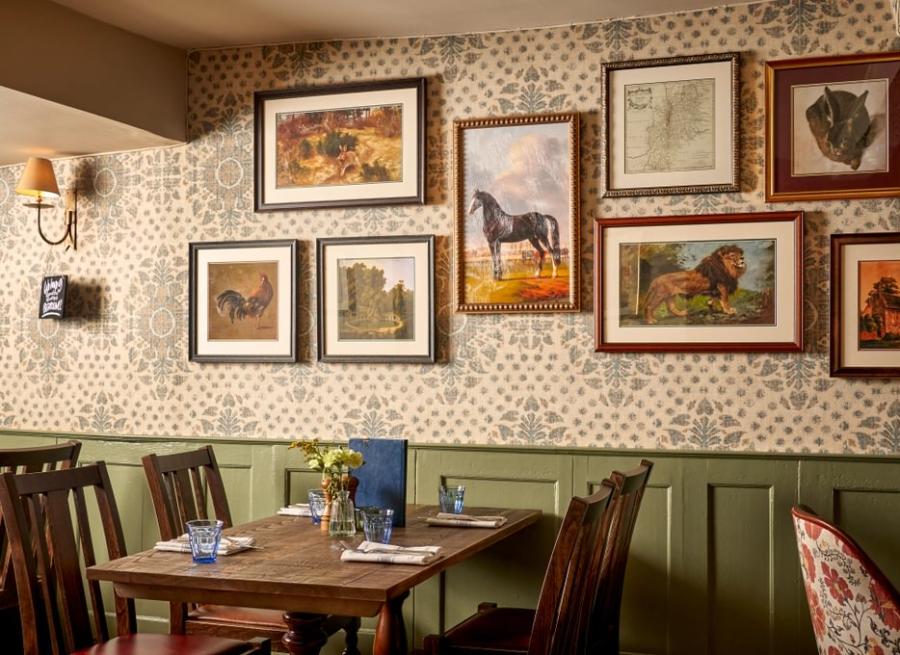 Inside the cosy restaurant at The Lion Inn. A wooden table set for four, surrounded by dark wooden chairs. The wall behind features patterned wallpaper and is decorated with a gallery of framed artwork, many depicting horses and animals in various styles and sizes. A small vase of yellow flowers sits on the table alongside menus, plates, and cutlery. Warm, ambient lighting creates an inviting, traditional atmosphere.